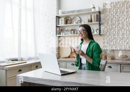 A young woman sits at home in the kitchen with a laptop and drinks morning coffee, holding a cup in her hands. Coffee break during working hours. Resting, looking dreamily out the window, smiling. Stock Photo