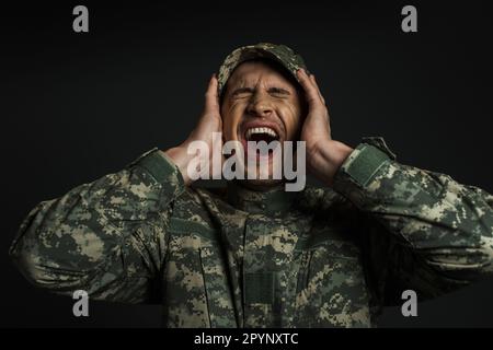 stressed soldier in camouflage uniform and cap screaming while suffering from ptsd isolated on black,stock image Stock Photo