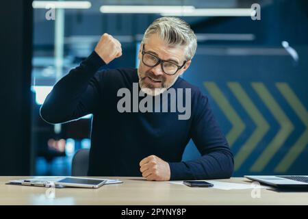 Portrait of an angry senior businessman sitting in the office in front of the camera and shouting, aggressively gesturing with his hands, pounding his fist on the table in an orderly manner. Stock Photo