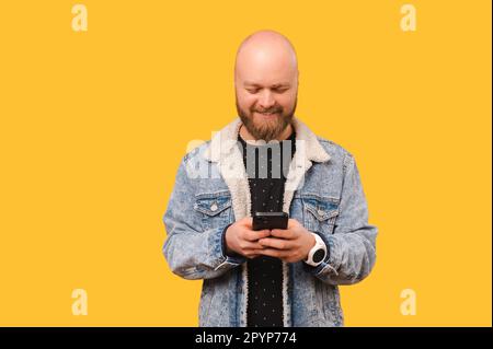 Handsome bearded bald man wearing a jeans jacket is texting and checking his phone in a yellow studio. Stock Photo