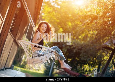 happy young female riding on macrame swing chair near country house outdoors Stock Photo