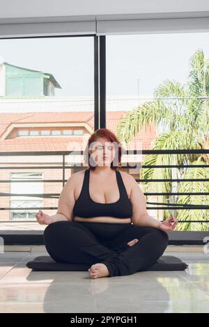 vertical image young plus size latin woman, doing yoga meditating with eyes closed in lotus position, indoors, wearing black sportswear. Stock Photo