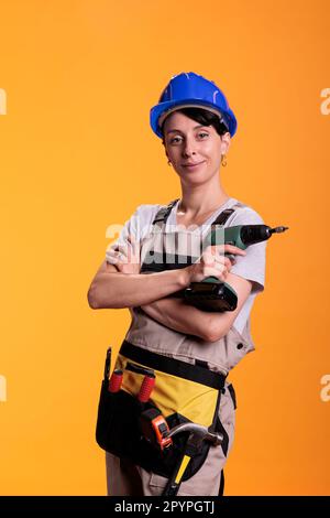 Young renovating worker posing on camera with screwing gun, using tool to drill nails and redecorate walls. Woman contractor wearing building uniform, indutrial engineering skill. Stock Photo