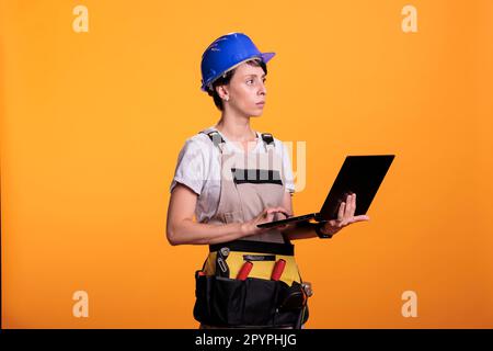 Focused construction worker holding wireless pc on camera, using laptop to find building materials or inspiration in studio. Woman builder reading online website on portable computer. Stock Photo