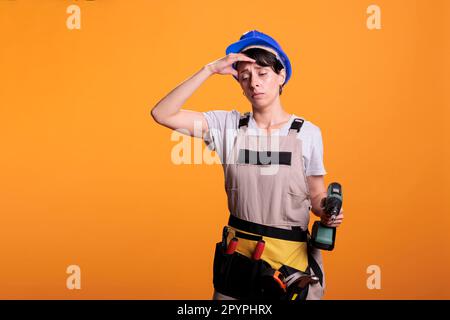 Tired young contractor with drilling gun feeling sleepy and fatigued, falling asleep at renovating project work. Female builder being exhausted with burnout and holding drill screw tool. Stock Photo
