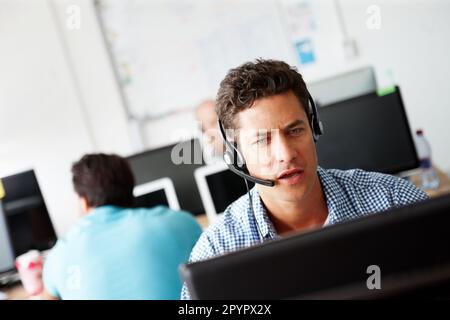 Making sense of the data. A handsome young sales assistant talking on his headset while working on a computer and colleagues working in the background Stock Photo
