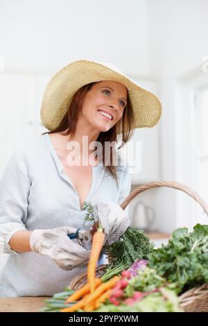 The simple life. A gorgeous woman, wearing a straw hat, cuts the stems off fresh vegetables in a basket on the kitchen counter. Stock Photo