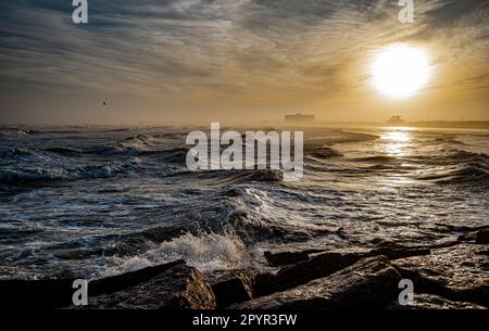 Waves roll onto a beach as the sun is low in the sky, in ominous dramatic seascape landscape. Stock Photo