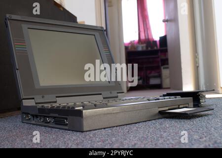 hands of a person taking a floppy disk and placing it inside an old laptop computer. The person is on the floor of a room Stock Photo