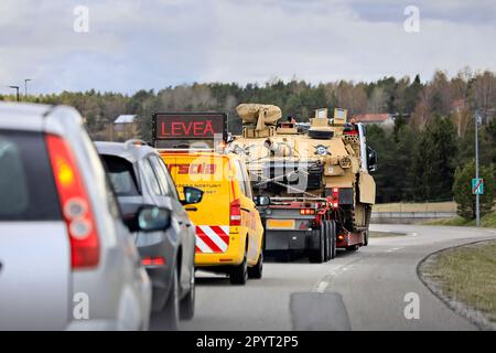 Truck transports American M1 Abrams military tank on low loader trailer on motorway ramp assisted by escort vehicle. Salo, Finland. April 28, 2023. Stock Photo