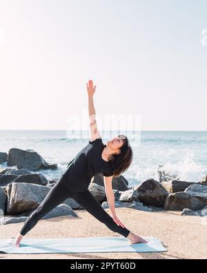 Slender female in sportswear standing barefoot on mat in Trikonasana while doing yoga on sandy beach against sea in summer Stock Photo