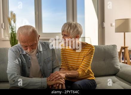 Mature woman comforting worried husband on sofa in living room by holding his hands Stock Photo