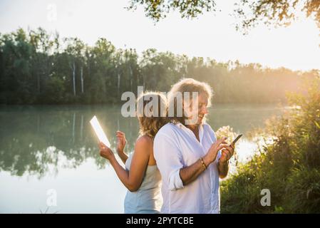 Couple at a river using smart phone and digital tablet, back to back Stock Photo