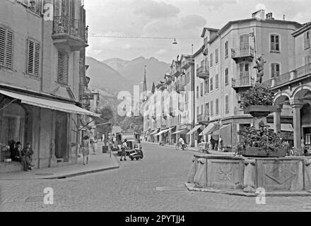 A street scene featuring a fountain in the Old Town, Chur, Graubunder, Switzerland in 1949. The water feature forms a ‘roundabout’ at the convergence of several streets in the town centre. It is capped by a sculpture of a bear holding a standard. Shops are open and shoppers are out and about. A drinks delivery truck is parked in the road. This is from an old amateur black and white 35mm negative – a vintage post-war photograph. Stock Photo