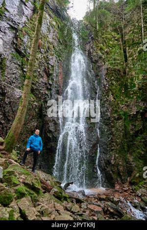 Tourist attraction near falls of Burgbach Waterfall. Man hiker in blue jacket standing on stone and looks at flow of falling water. Waterfall Stock Photo