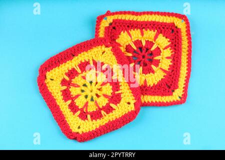 Crochet granny squares. Two red and yellow squares with circular pattern in the centre changing to square near the edges. Aqua blue background. Stock Photo