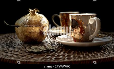 A luxurious gold-colored vase situated to the side of two white coffee cups placed on a decorative plate Stock Photo