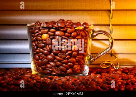 Glass coffee cup filled with raw roast coffee beans set against a kitchen window blind Stock Photo