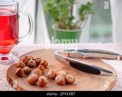 A glass of red berry tea and hazelnuts with nutcracker lying on a wooden board on a cozy orange warm cloth. Window with curtain lace in the background Stock Photo