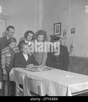 Undated photo. In 1950, a family is watching a photo album with the photos of their fallen soldier family member in a living room in Berlin, presumably West Berlin.  The parents are sitting in the front. Behind them, children and a granddaughter. On the right, the father wears the uniform of the Deutsche Reichsbahn (German State Railroad). Stock Photo