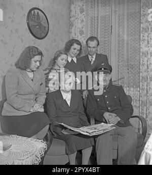 Undated photo. In 1950, a family in a living room in Berlin is watching a photo album with photos of a relative who have fallen as a soldier, presumably West Berlin.  The parents are sitting in the front. Behind them, children and a granddaughter. On the right, the father wears the uniform of the Deutsche Reichsbahn (German State Railroad). Stock Photo