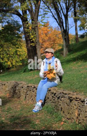 In the photo, a young woman with a bouquet of yellow leaves sits and dreams about something in the autumn park. Stock Photo