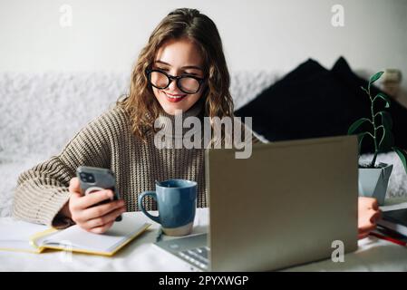 SMM specialist working from home at kitchen table, using smartphone to select blogger for product promotion in social media, in front of laptop Stock Photo