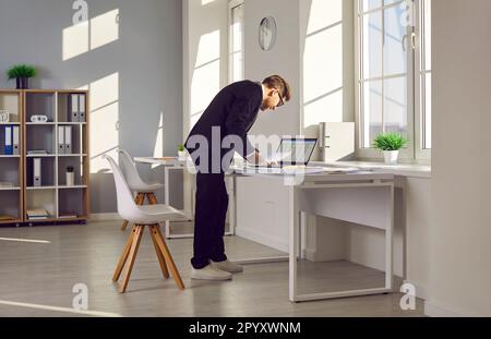 Accountant standing by his office desk, doing some paperwork, and using a laptop computer Stock Photo
