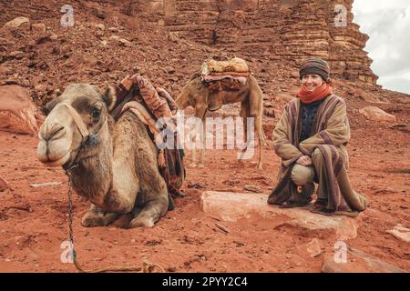 Young woman in traditional Bedouin coat - bisht - and headscarf crouching next to camel laying on red desert ground, smiling, rocky wall background Stock Photo