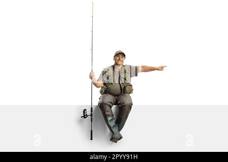 Cheerful mature fisherman with a fishing rod sitting on a blank panel and pointing to the side isolated on white background Stock Photo