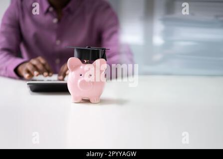 Businessperson's Hand Using Calculator Beside Piggybank With Graduation Hat Stock Photo