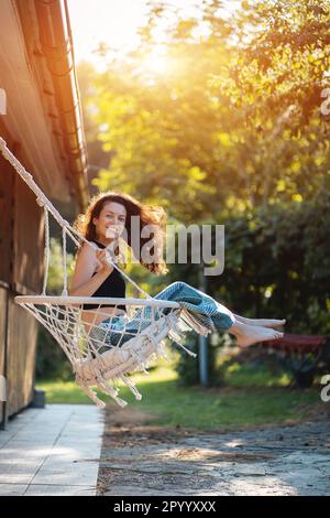 happy young female riding on macrame swing chair near country house outdoors Stock Photo