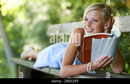 Doing some light reading. Pretty young woman lying on a bench and reading her book outdoors. Stock Photo