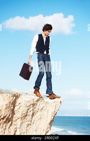 Living on the edge. Young semi-formal businessman stepping towards the edge of a cliff overlooking the ocean. Stock Photo