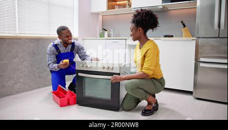 Smiling Repairman Repairing Oven On Kitchen Worktop In Front Of Woman Stock Photo