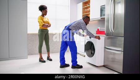 Woman Looking At Young Male Worker With Toolbox Repairing Washing Machine In Kitchen Room Stock Photo