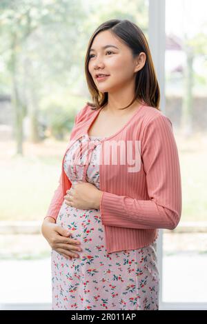 Happy young pregnant Asian woman in maternity clothes standing in her living room holding her belly thinking of her unborn child Stock Photo