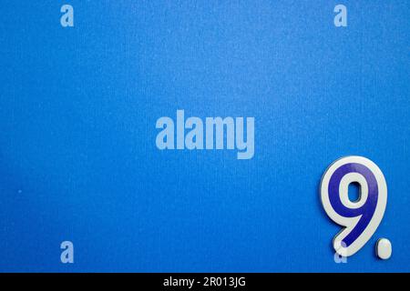 The number 9 in light blue and white color, photographed from above, placed on the edge of a blue background. Stock Photo