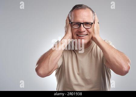 Hear no evil. a mature man standing against a grey background in the studio and covering his ears with his hands. Stock Photo