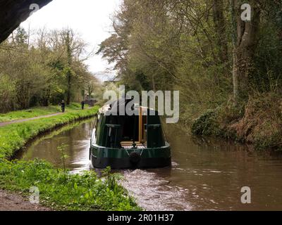 Helmsman in green narrowboat steering through bridge on Monmouthshire and Brecon Canal with two cyclists riding along towpath Talybont-on-Usk Powys Mi Stock Photo
