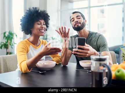 Social media always have us debating something. a young couple looking at something on a cellphone while having breakfast at home. Stock Photo