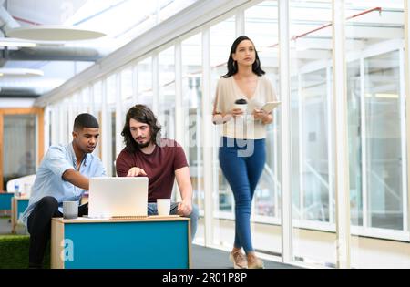 Taking a moment to sit with their plans. two businessmen working together on a laptop in an office with their colleague walking in the background. Stock Photo