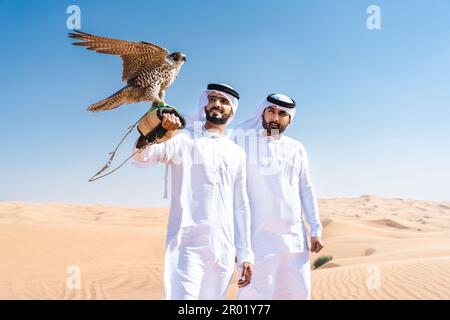 Two middle-eastern men wearing traditional emirati arab kandura bonding in the desert and holding a falcon bird - Arabian muslim friends meeting at th Stock Photo