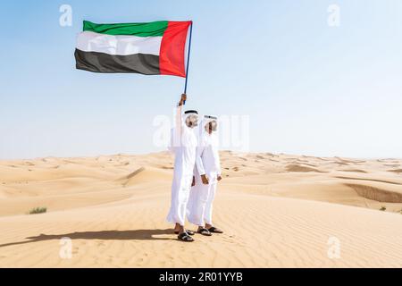 Two middle-eastern men wearing traditional emirati arab kandura bonding in the desert and holding emirati flag -  Arabian muslim friends meeting at th Stock Photo