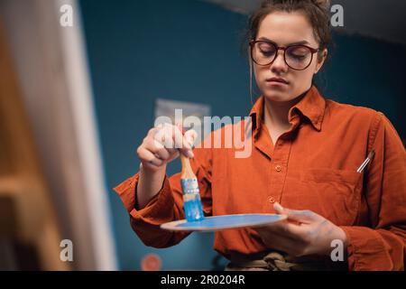 inspirational concentrated latino woman in apron draws pictures with paints, standing in modern studio or living room. Experienced female artist Stock Photo