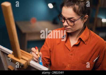 inspirational concentrated latino woman in apron draws pictures with paints on easel, standing in modern studio or living room. Happy experienced Stock Photo