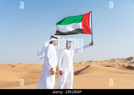 Two middle-eastern men wearing traditional emirati arab kandura bonding in the desert and holiding the UAE flag to celebrate national day - Arabian mu Stock Photo