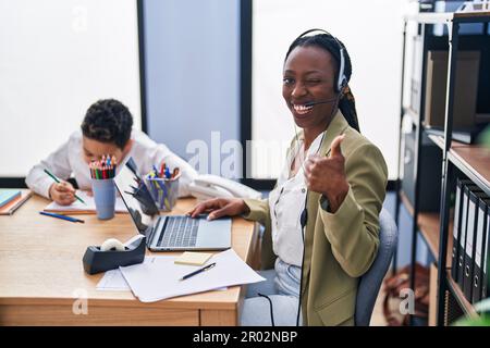 Young mother wearing call center agent headset smiling happy and positive, thumb up doing excellent and approval sign Stock Photo