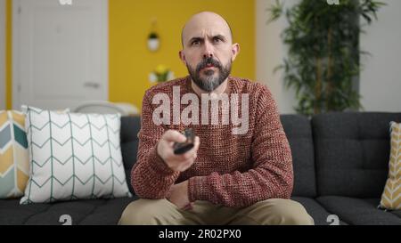 Young bald man watching tv sitting on sofa with boring expression at ...