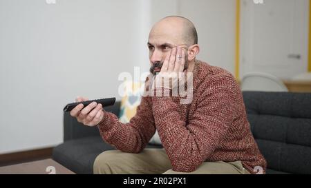 Young bald man watching tv sitting on sofa with boring expression at ...